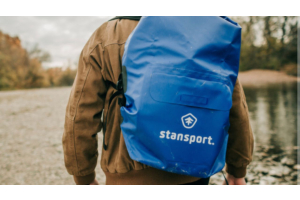 Person carrying blue Stansport waterproof dry bag backpack by river.
